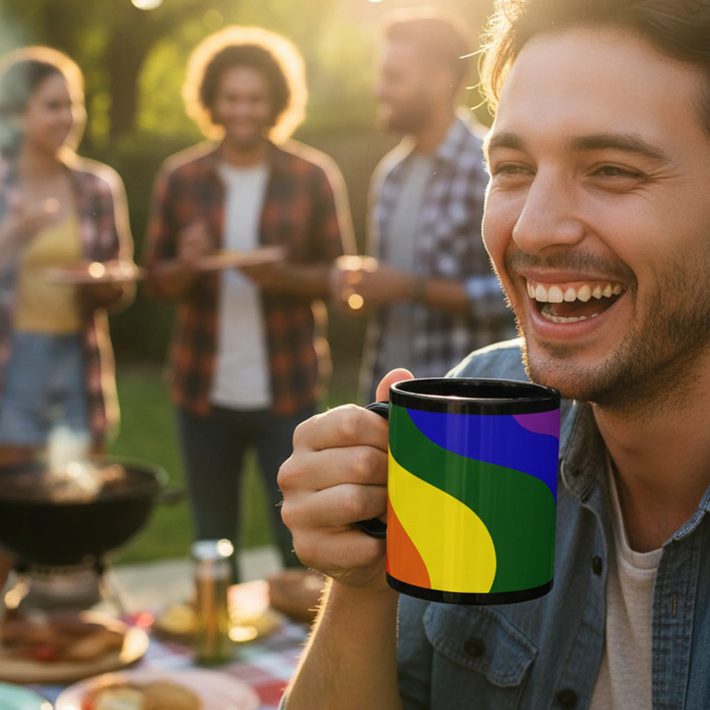Man smiling outdoors holding a black FREEBRD rainbow wave ceramic mug with rainbow pride design in yellow green blue and orange.