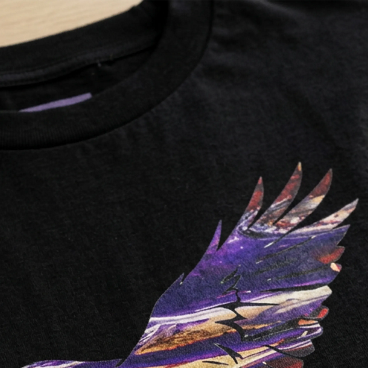 Close-up of purple, gold, gray, and white Tribal Wave artwork inside a flying raven graphic on a black T-shirt.
