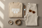A summer lifestyle bird’s-eye lay of the Freebrd Logo Classic T-Shirt in sand that has a white Freebrd Logo on the chest, brown shade sunglasses, a classic watch, cream jeans, belt and beads on a small tea plate.