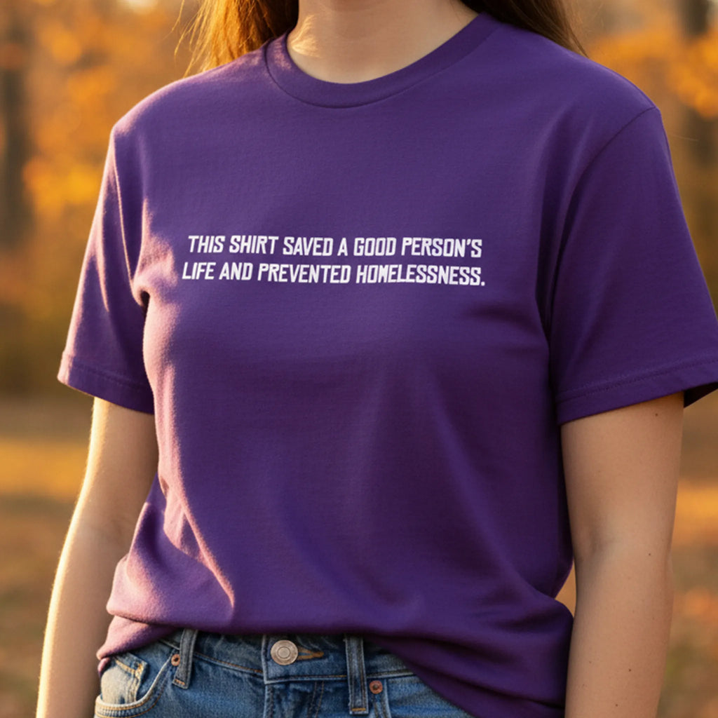 Purple cotton t-shirt with white block text reading This shirt saved a good person's life and prevented homelessness, worn with blue jeans, front view on person outdoors.