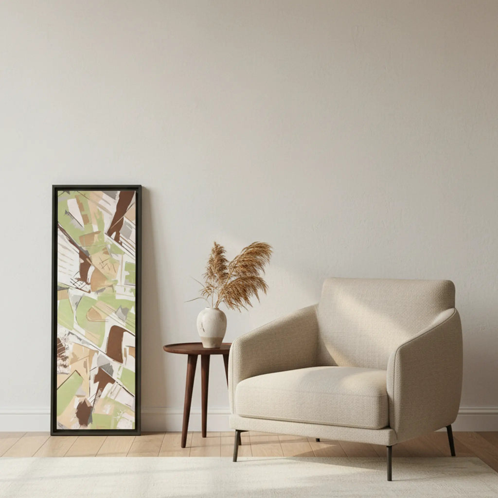 Freebrd "Slim Sage" framed abstract canvas art, 12x36 vertical, 1.25" thick, fully framed in black wood, leaning against modern interior wall beside accent sofa chair — size reference in sage green, taupe, and muted brown.