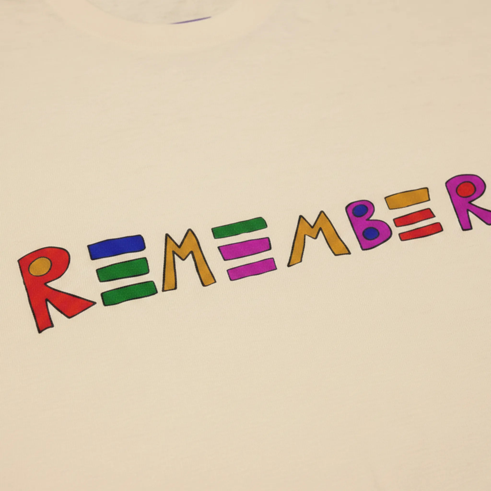 Classic fit crew neck tee, sand, close-up texture showing cotton fabric weave and hand-drawn multicolor REMEMBER graphic in red, blue, green, orange, purple, and pink.