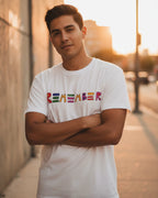 Man wearing a white spread love daily postive message t-shirt with 'REMEMBER' printed in colorful letters, standing on a street with a blurred background.