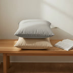 Bird's eye view of stardust grey and natural jute beige 18x18 faux suede throw pillows stacked on oak bench for a size reference.