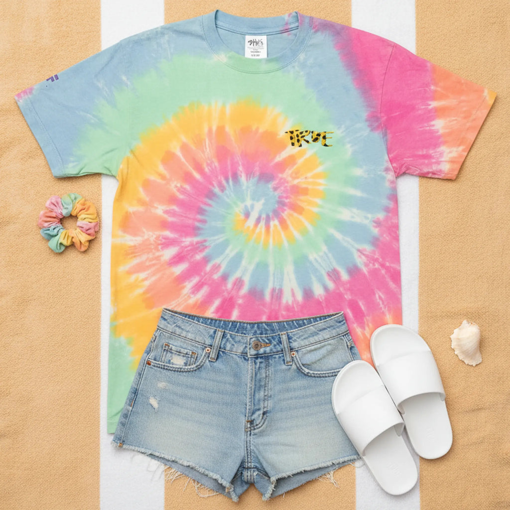 A bird's eye flat lay of the pastel sherbet tie-dye cheetah embroidered tee styled in a feminine summer outfit with denim cutoff shorts, white slide sandals, a scrunchie, and a seashell on a sandy beach towel surface.
