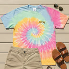 A bird's eye flat lay of the pastel sherbet tie-dye cheetah embroidered tee styled in a masculine summer outfit with khaki shorts, brown leather sandals, a woven bracelet, and sunglasses on a light wood deck surface.