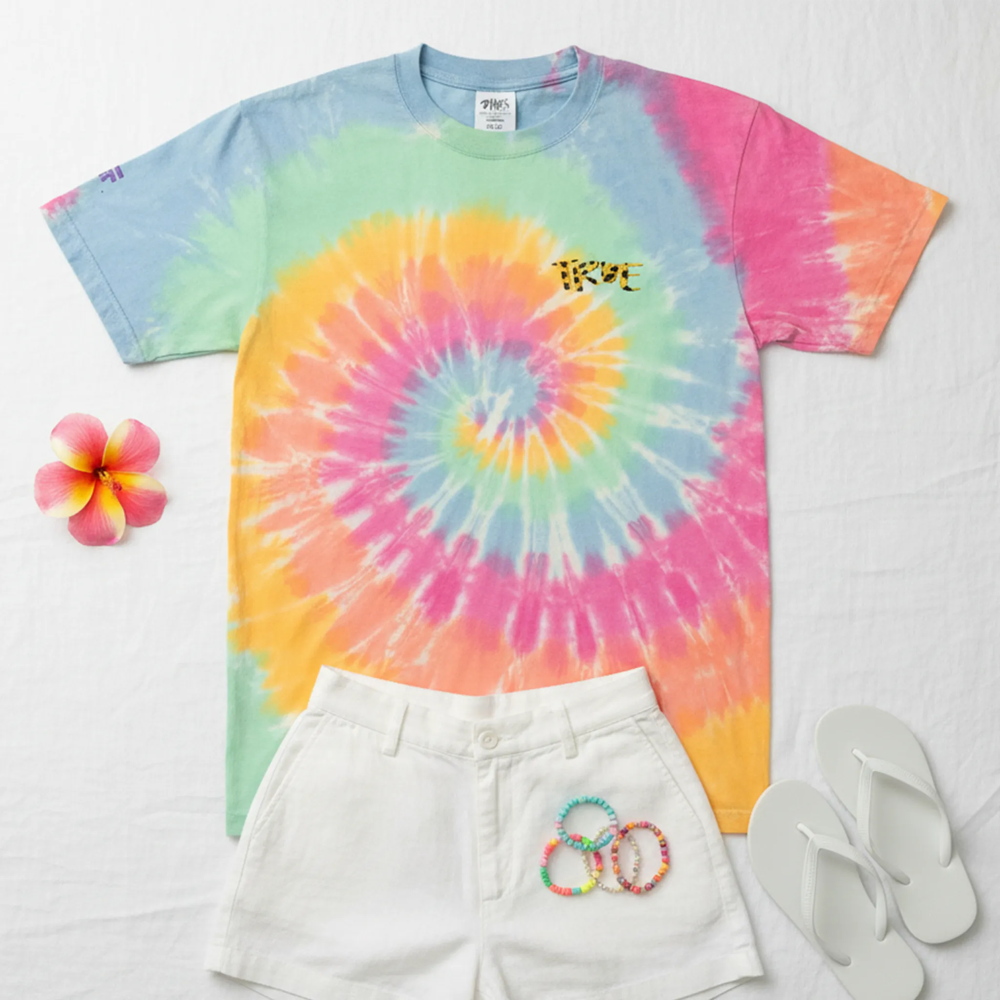 A bird's eye flat lay of the pastel sherbet tie-dye cheetah embroidered tee styled in a unisex summer outfit with white linen shorts, colorful beaded bracelets, white flip flops, and a tropical flower on a white linen surface.