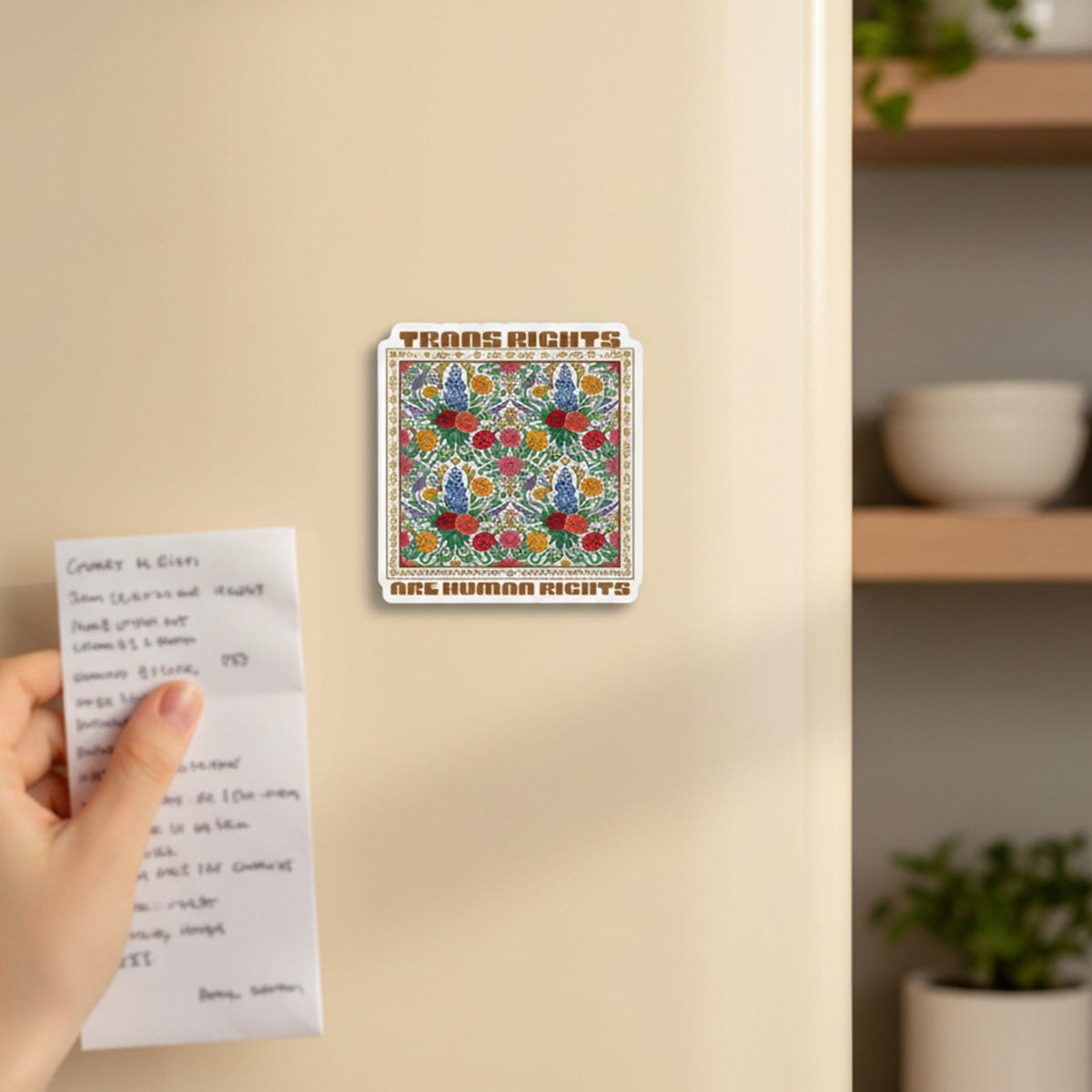 A rectangular Trans Rights Are Human Rights refrigerator magnet with a colorful floral pattern, shown attached to a beige refrigerator door. A hand is holding a small handwritten grocery list in the foreground as a size reference.