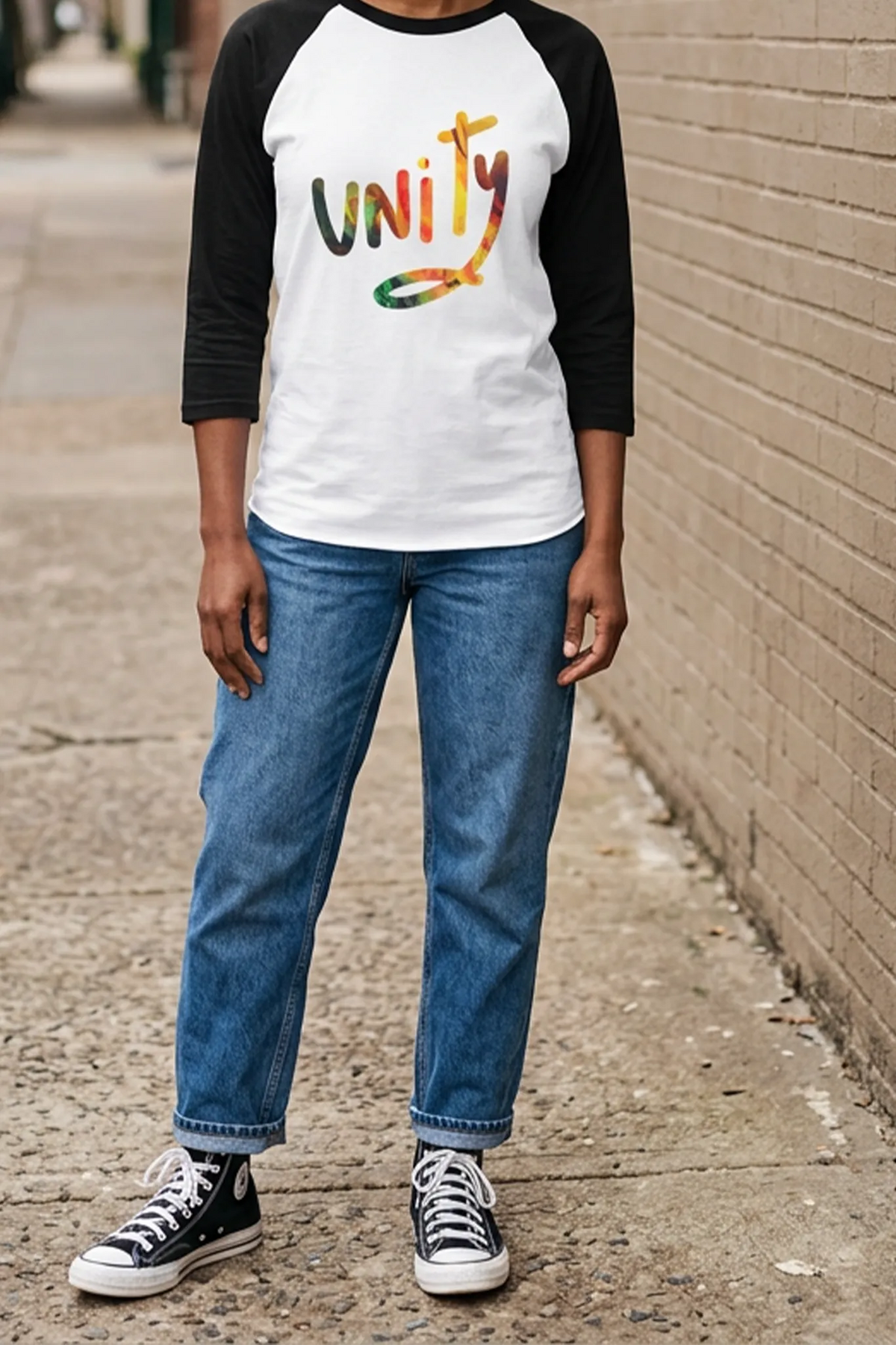 Woman wearing a white raglan with black three-quarter sleeves and multicolored unity graphic, cuffed blue jeans, and black high-top sneakers beside a brick wall.