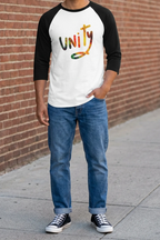 Man wearing a white raglan with black three-quarter sleeves and multicolored unity graphic, cuffed blue jeans, and black high-top sneakers beside a brick wall.