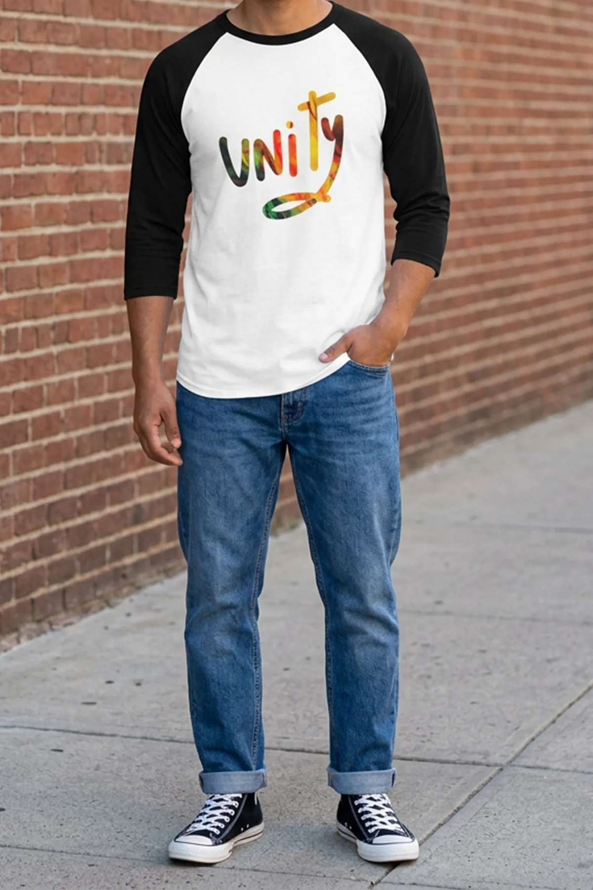 Man wearing a white raglan with black three-quarter sleeves and multicolored unity graphic, cuffed blue jeans, and black high-top sneakers beside a brick wall.