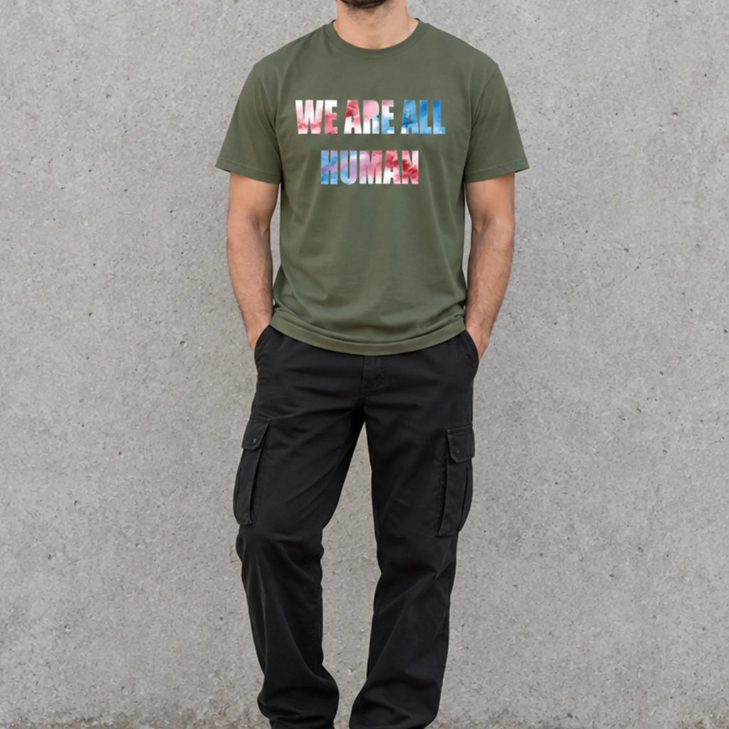 Model wearing a military green ‘WE ARE ALL HUMAN’ graphic T-shirt with black cargo pants and black sneakers, standing against a concrete wall.
