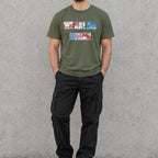 Model wearing a military green ‘WE ARE ALL HUMAN’ graphic T-shirt with black cargo pants and black sneakers, standing against a concrete wall.