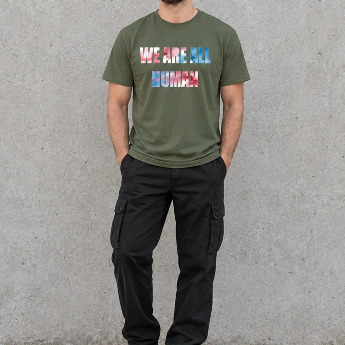 Model wearing a military green ‘WE ARE ALL HUMAN’ graphic T-shirt with black cargo pants and black sneakers, standing against a concrete wall.