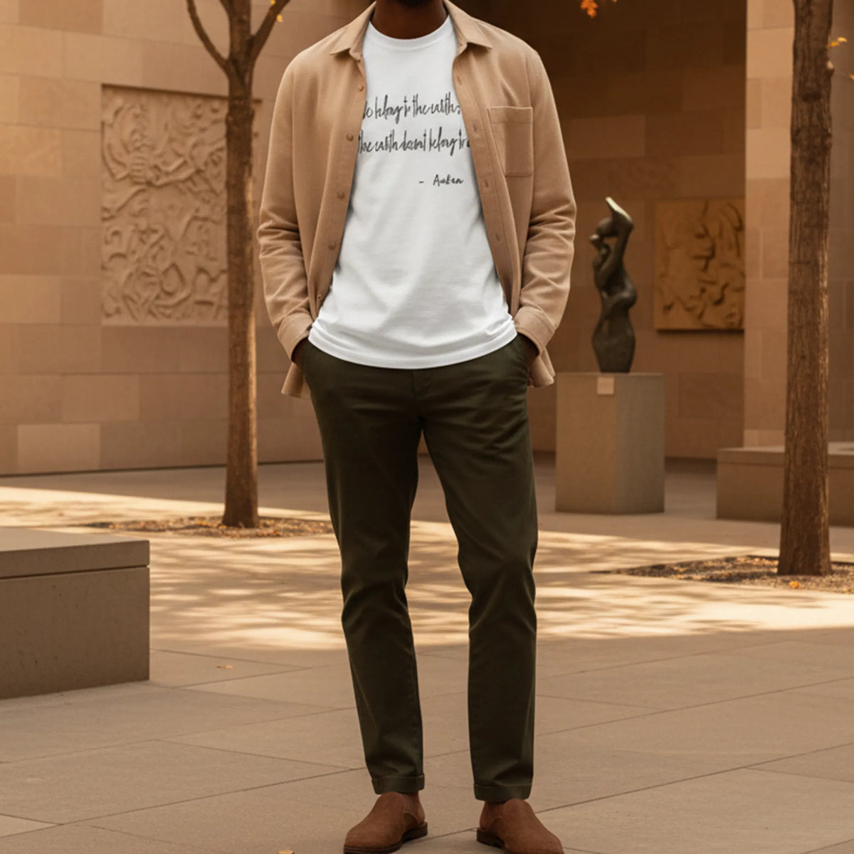 Adult model wearing a white tee with black handwritten chest lettering under an oatmeal overshirt in a sunlit autumn art museum courtyard.